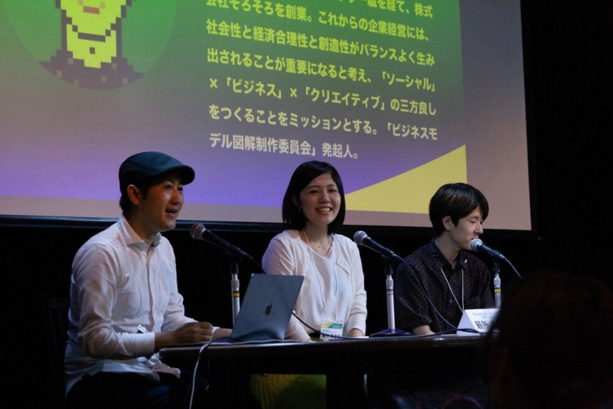 壇上での鼎談。3人がマイクの前に並び、スクリーンには近藤哲朗の紹介文が映っている。