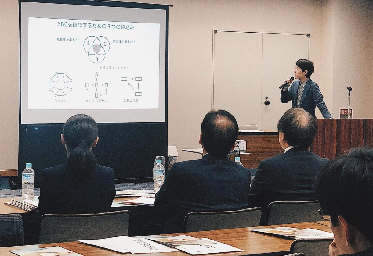 壇上で話す近藤哲朗。スクリーンには「SBCを確認するための3つの枠組み」として、社会性・経済性・創造性の3つの円と、八方よし・ビジネスモデル・逆説の構造の3つの図が映っている。