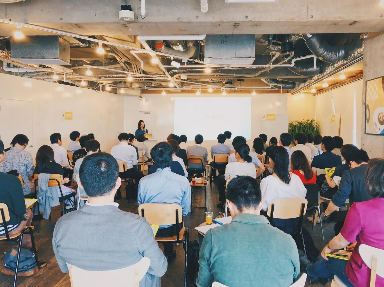 イベント会場。椅子を並べた部屋で、参加者が前方のスクリーンを見ている。