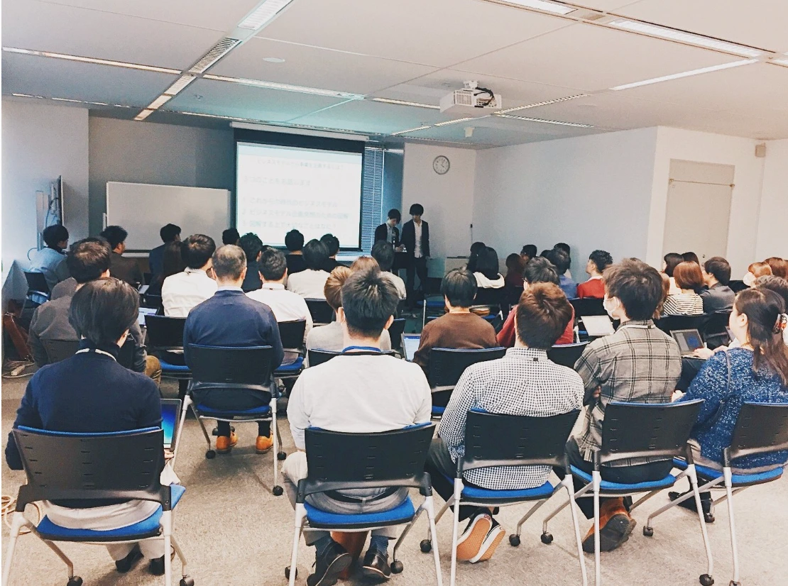 講演会場を後ろから写したところ。椅子が並び、参加者がスクリーンを見ている。